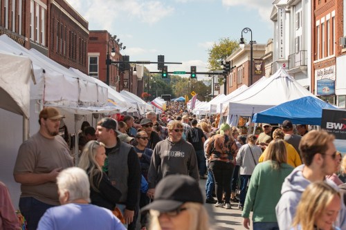 A bustling street fair with crowds of people walking between vendor tents under a clear blue sky.