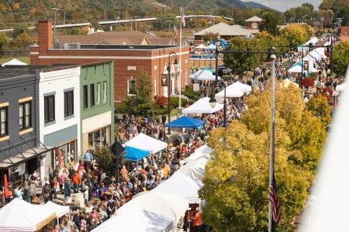 A bustling street festival with crowds, tents, and colorful autumn trees in a small town.