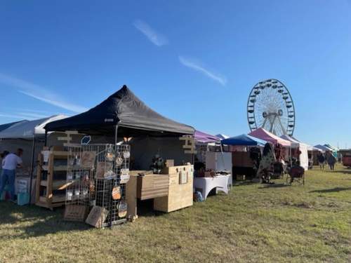 A vibrant outdoor market with various vendor tents and a large Ferris wheel in the background under a clear blue sky.