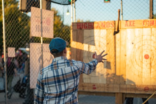 A person in a plaid shirt throws an axe at a wooden target in a fenced area during an outdoor event.