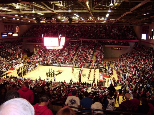 A packed basketball arena with fans in red, watching a game with players on the court and a large scoreboard overhead.