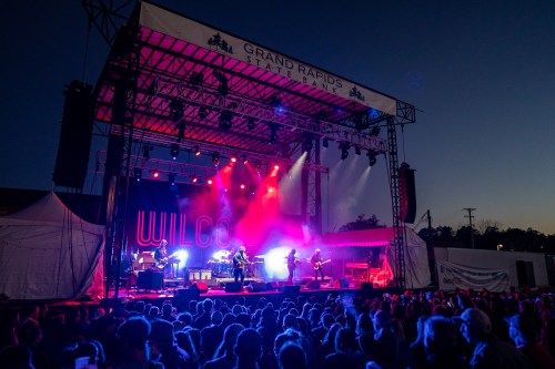 A band performs on stage with colorful lights in front of a large crowd at a music festival during twilight.