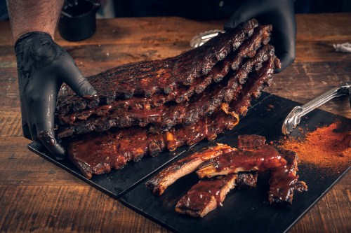 A person in black gloves holds a stack of glazed ribs on a wooden table, with some ribs laid out beside them.