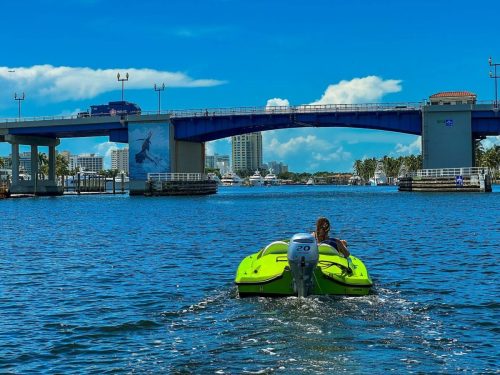 A person rides a bright green jet ski under a blue bridge on a sunny day, with buildings and boats in the background.