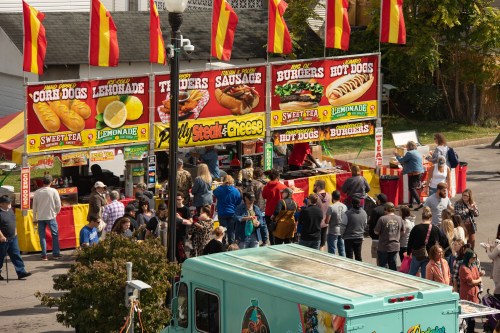 A busy food festival scene with people lining up at colorful food stalls offering corn dogs, burgers, and lemonade.