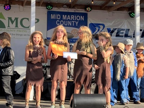 Four girls in cavegirl costumes stand on stage, smiling and holding a paper, with a crowd and banners in the background.
