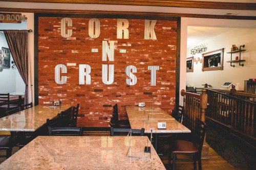 Interior of a restaurant with a wooden wall displaying "CORK -N- CRUST" and empty tables with clear partitions.