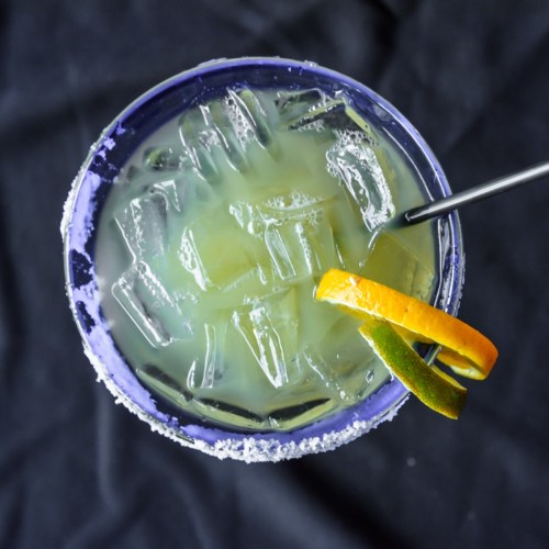 A top-down view of a colorful cocktail with ice, garnished with orange and lime slices, in a salt-rimmed glass.