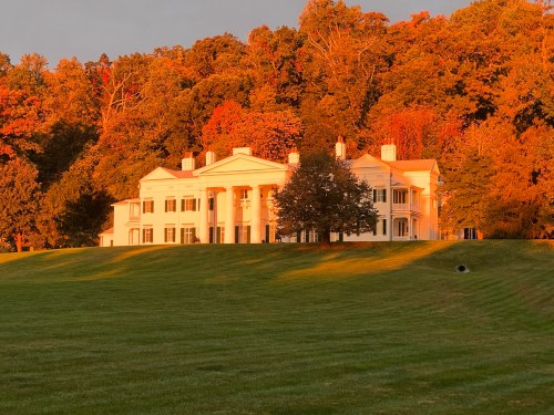 A grand white mansion with columns, surrounded by vibrant autumn foliage and a green lawn, illuminated by sunset.