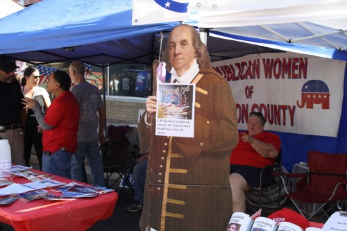 A cardboard cutout of Benjamin Franklin stands at a booth for Republican Women of Washington County, with people nearby.