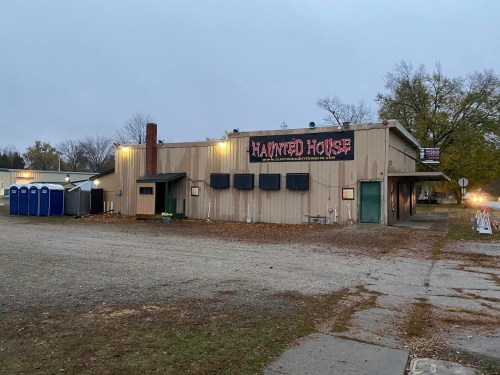 A spooky haunted house building with a sign, surrounded by trees and portable restrooms in a gravel lot.