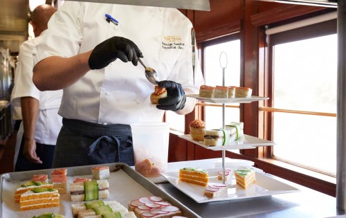 A chef in a white uniform prepares colorful sushi and desserts on a tiered stand in a train dining car.
