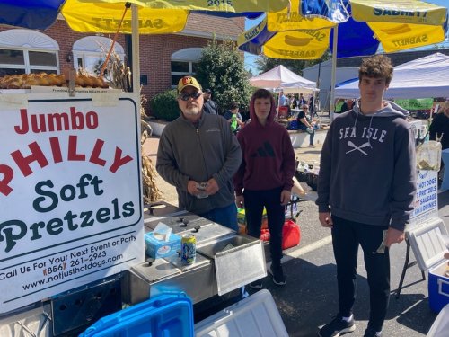 Three people stand at a food stall with a sign for "Jumbo Philly Soft Pretzels" at an outdoor event.