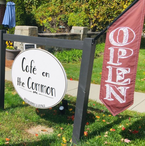 Sign for "Café on the Common" with an "Open" banner, surrounded by greenery and autumn leaves.