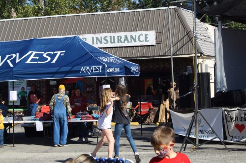 A lively outdoor event with a blue tent, people browsing merchandise, and children playing in front of a storefront.