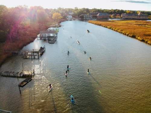 Aerial view of a river with people paddleboarding, surrounded by trees and buildings under a sunny sky.