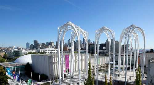 A modern architectural structure with white arches, overlooking a city skyline under a clear blue sky.