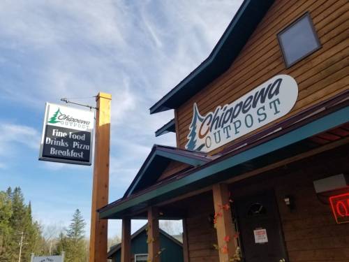 Sign for Chippewa Outpost featuring "Fine Food, Drinks, Pizza, Breakfast" on a wooden building with a blue sky backdrop.