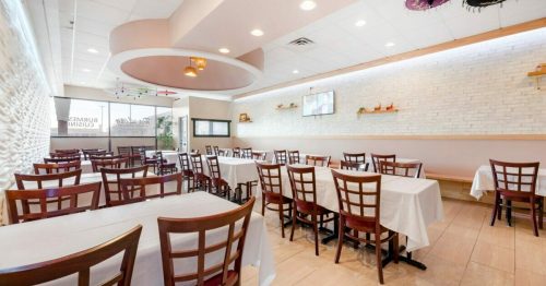 Interior of a restaurant featuring empty tables with white tablecloths, bright lighting, and decorative wall elements.