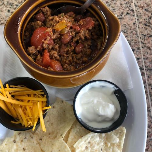 A bowl of chili with ground meat, beans, and tomatoes, served with cheese, sour cream, and tortilla chips on the side.