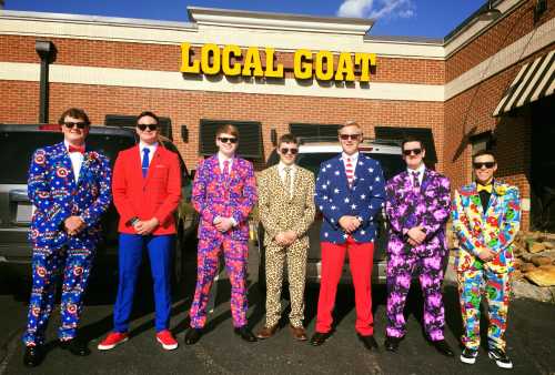 A group of seven men in colorful, patterned suits stand outside a restaurant called "Local Goat."