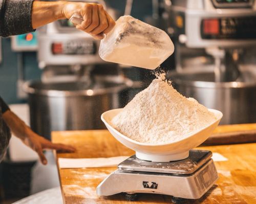 A hand pours flour from a scoop into a bowl on a digital scale, with kitchen equipment in the background.