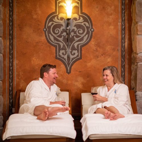 A man and woman in white robes relax on lounge chairs, enjoying wine and laughing in a cozy spa setting.
