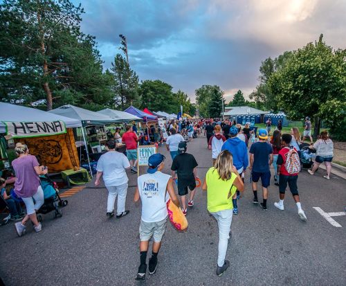 A lively outdoor festival scene with people walking among vendor tents and colorful decorations under a cloudy sky.