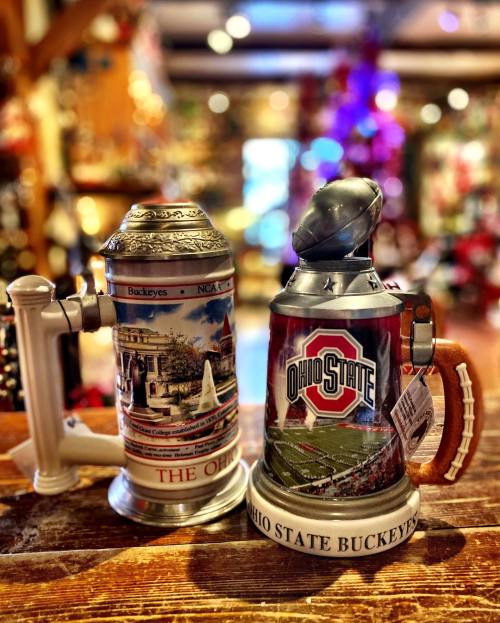 Two decorative steins featuring Ohio State Buckeyes designs, displayed on a wooden surface with festive background decor.