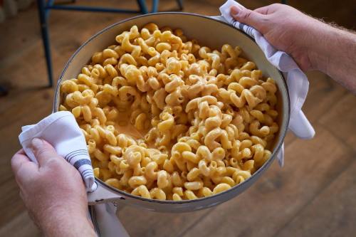 A person holds a large pot of creamy, cheesy pasta, with curly noodles, using white towels.