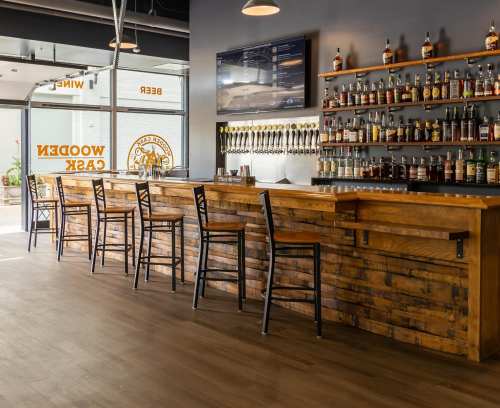 A wooden bar with high stools, shelves of liquor, and a TV in a modern, well-lit space.