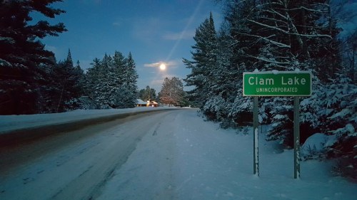 A snowy road leads to a sign for Clam Lake, illuminated by a bright moon in a serene winter landscape.