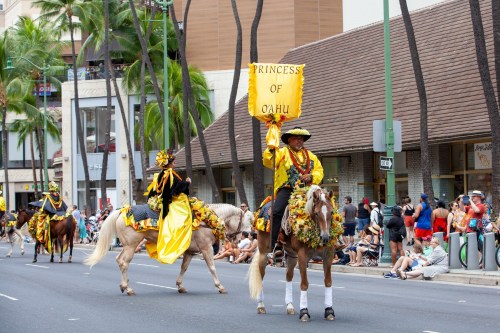 A parade scene featuring riders on horses, one holding a "Princess of Oahu" banner, with spectators along the street.