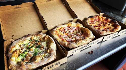 Three pizzas in cardboard boxes: one with vegetables, one with ham, and one with pepperoni, on a countertop.