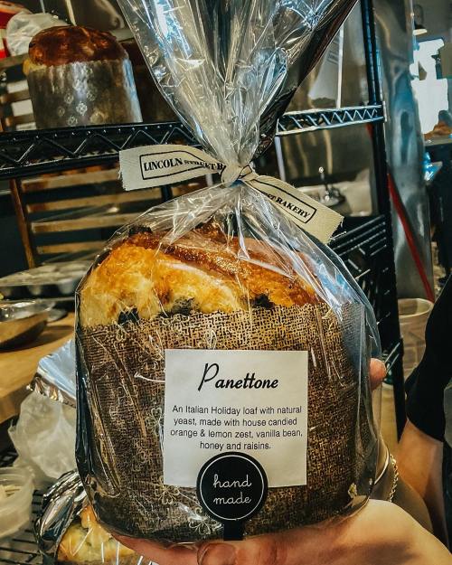 A wrapped panettone loaf with a label, showcasing its ingredients: orange, lemon zest, vanilla, honey, and raisins.