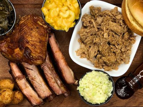A wooden platter featuring pulled pork, ribs, macaroni and cheese, collard greens, coleslaw, and a bun.