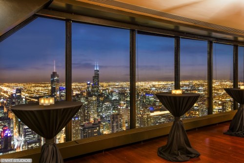 Elegant event space with tall tables overlooking a stunning city skyline at night, filled with lights.