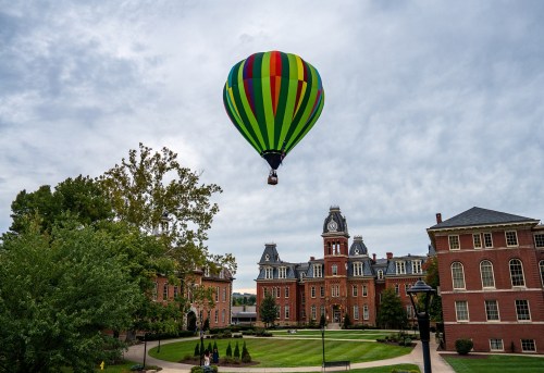 A colorful hot air balloon floats above a historic campus with brick buildings and green trees under a cloudy sky.