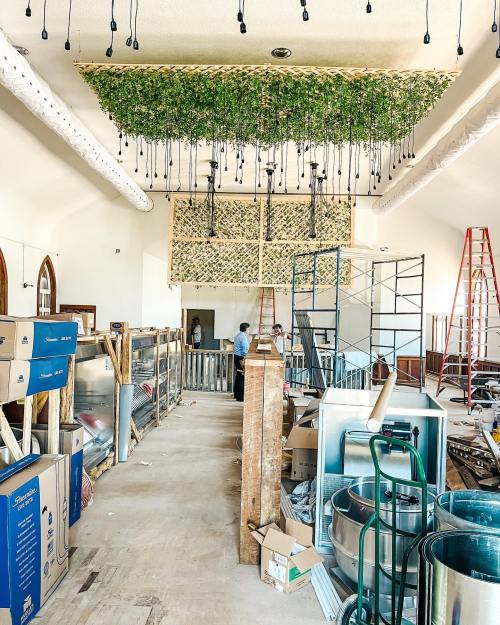 Interior of a construction site with hanging greenery, scaffolding, and workers amidst unfinished decor and materials.
