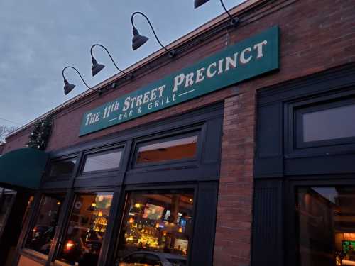 Sign for "The 11th Street Precinct Bar & Grill" on a brick building, with warm lights visible through the windows.