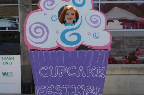 A person peeks through a cupcake cutout at a festival, surrounded by colorful decorations and flowers.