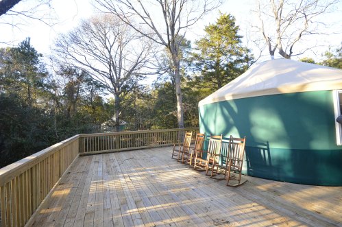 A wooden deck with rocking chairs beside a green yurt, surrounded by trees in a sunny outdoor setting.