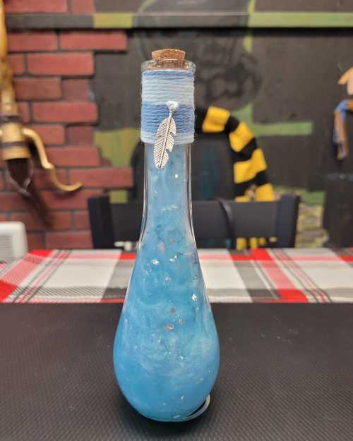 A decorative glass bottle with a cork stopper, filled with blue liquid and adorned with a silver feather charm.