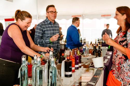 A lively wine tasting event with people sampling drinks and engaging at a table filled with various bottles.