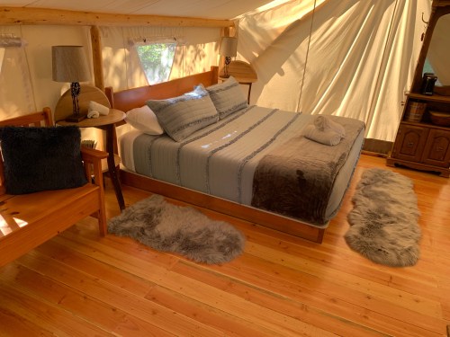 Cozy tent interior with a double bed, wooden furniture, and soft rugs on a wooden floor, illuminated by natural light.