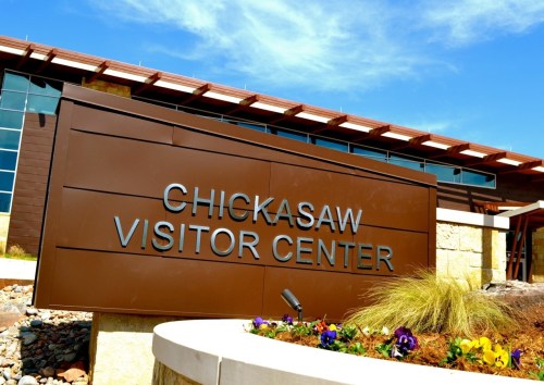Sign for the Chickasaw Visitor Center, featuring modern architecture and colorful flowers in the foreground.