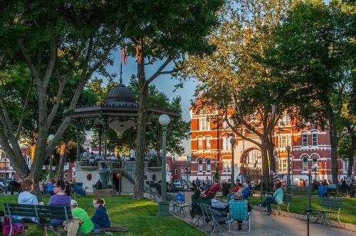 A park scene with a gazebo, people sitting on the grass, and a historic building in the background, surrounded by trees.