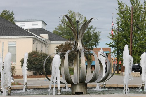 A modern metal fountain shaped like a flower, with water jets spraying around it in a public square.