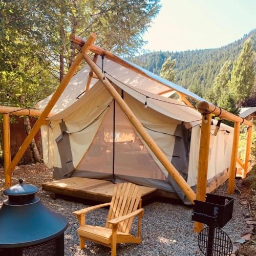 A cozy tent with wooden supports, set in a forested area, featuring a deck and an outdoor seating area.