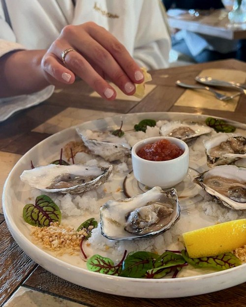 A hand squeezing lemon over a plate of oysters on ice, garnished with sauce and herbs.